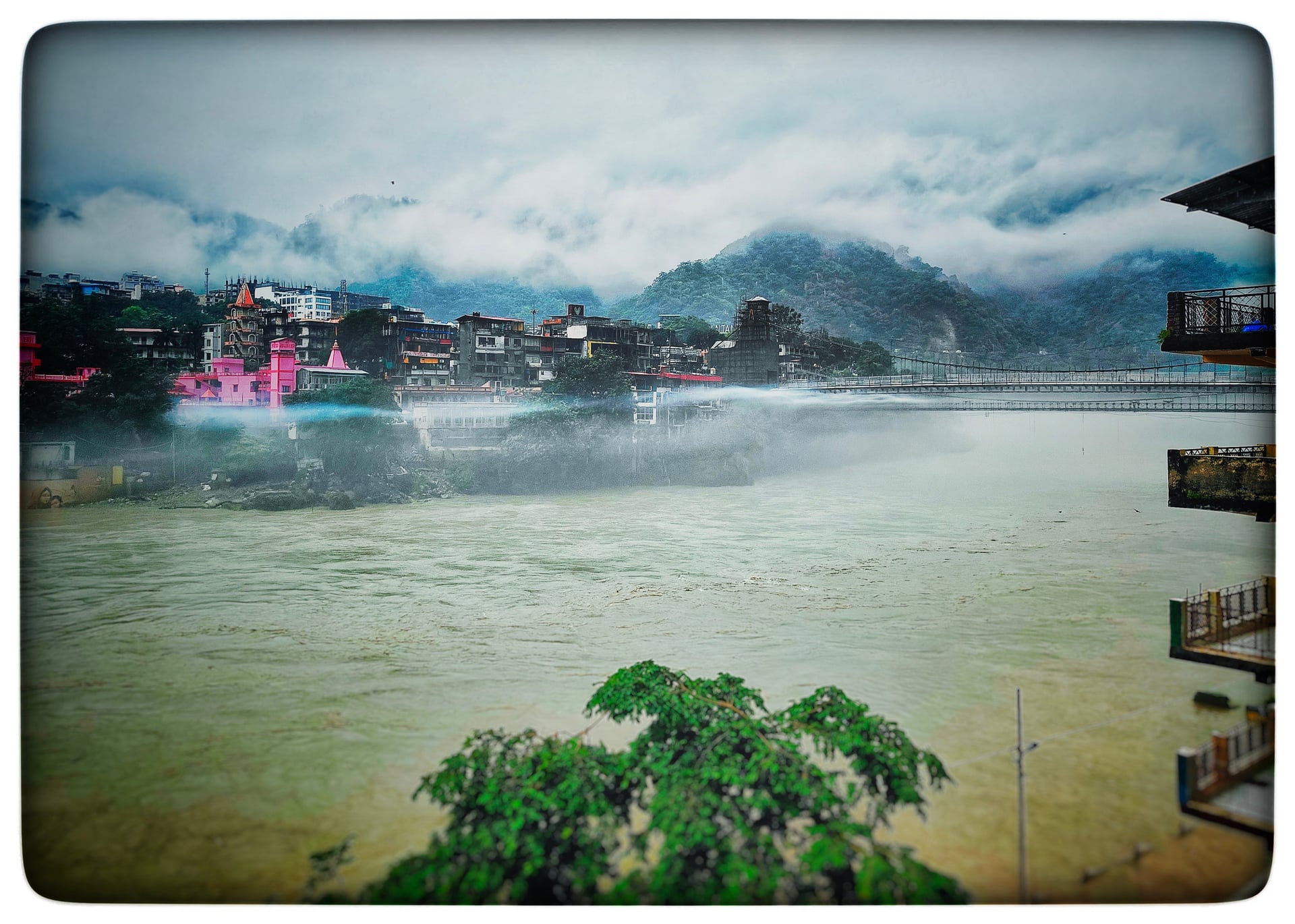 Panoramic view of the misty Ganges River flowing through Rishikesh, with vibrant buildings and a bridge under cloud-covered mountains.