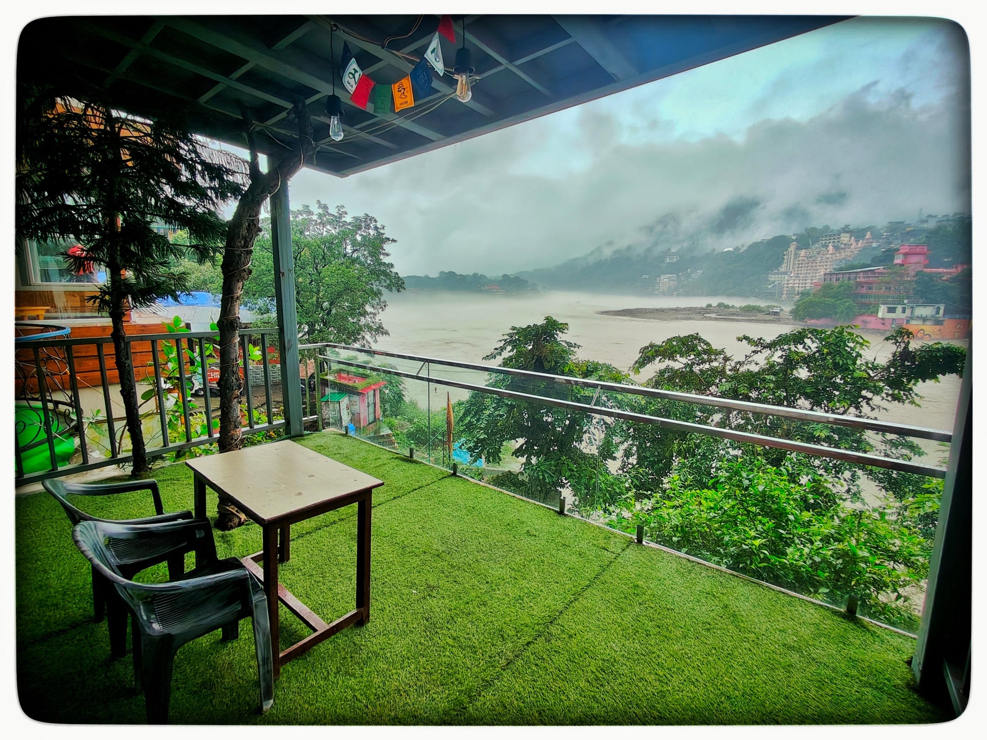 Hotel balcony with green artificial turf, a small table, two chairs, and panoramic views of the misty Ganges River and distant mountains.