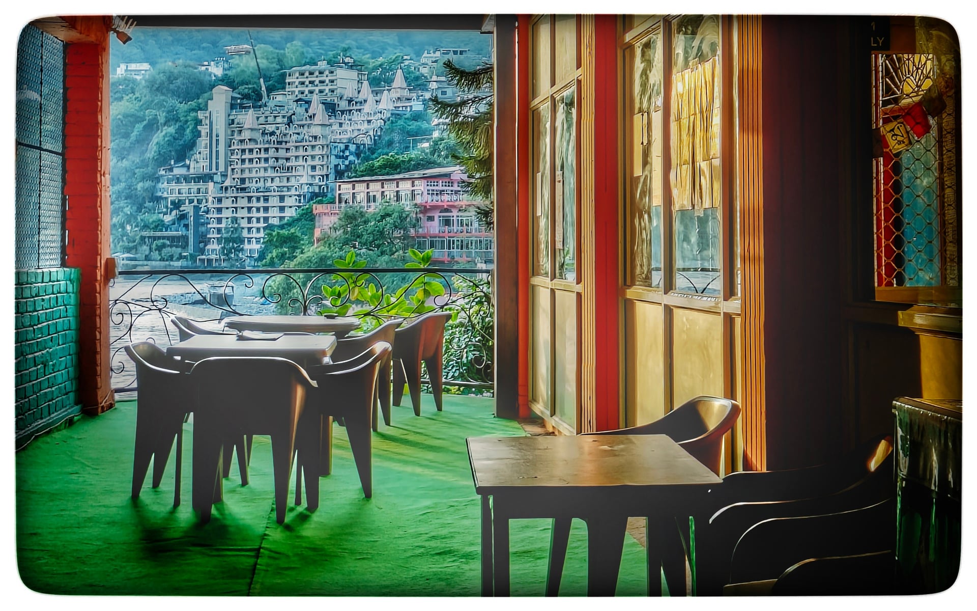 Outdoor seating area on a hotel terrace with a vibrant green floor, overlooking the Ganga River and hillside buildings.