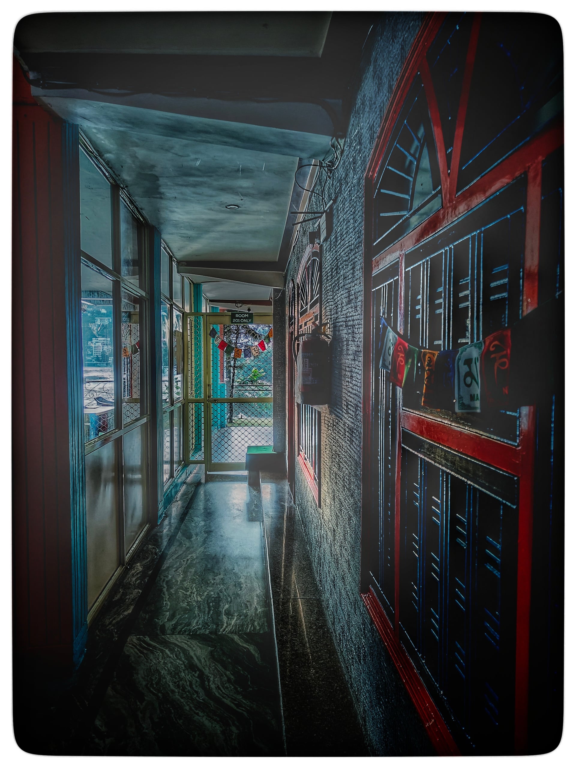 Narrow hotel corridor with dark marble floor, windows on left, and red-framed wall with prayer flags on right leading to an exit.