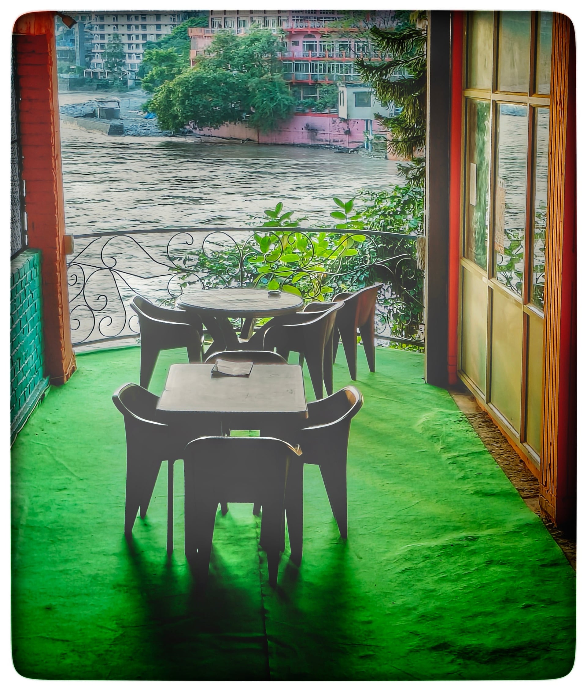 Outdoor terrace with simple tables and chairs, featuring a vibrant green floor and overlooking the wide Ganges River and distant buildings.