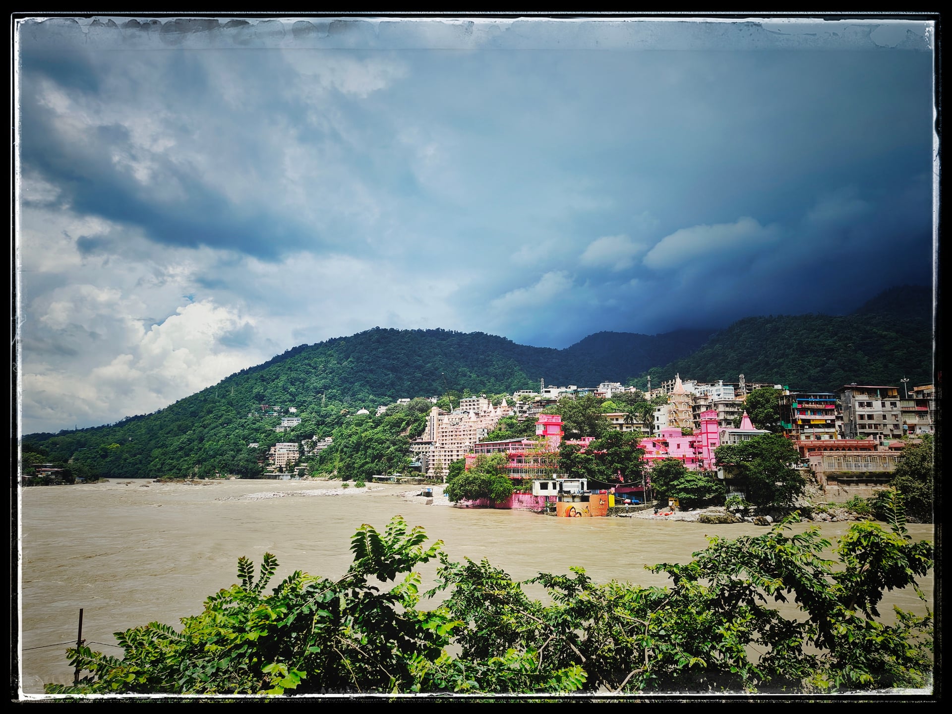 Panoramic view of the Ganges River, colorful buildings of Rishikesh, lush green hills, and dramatic cloudy sky.