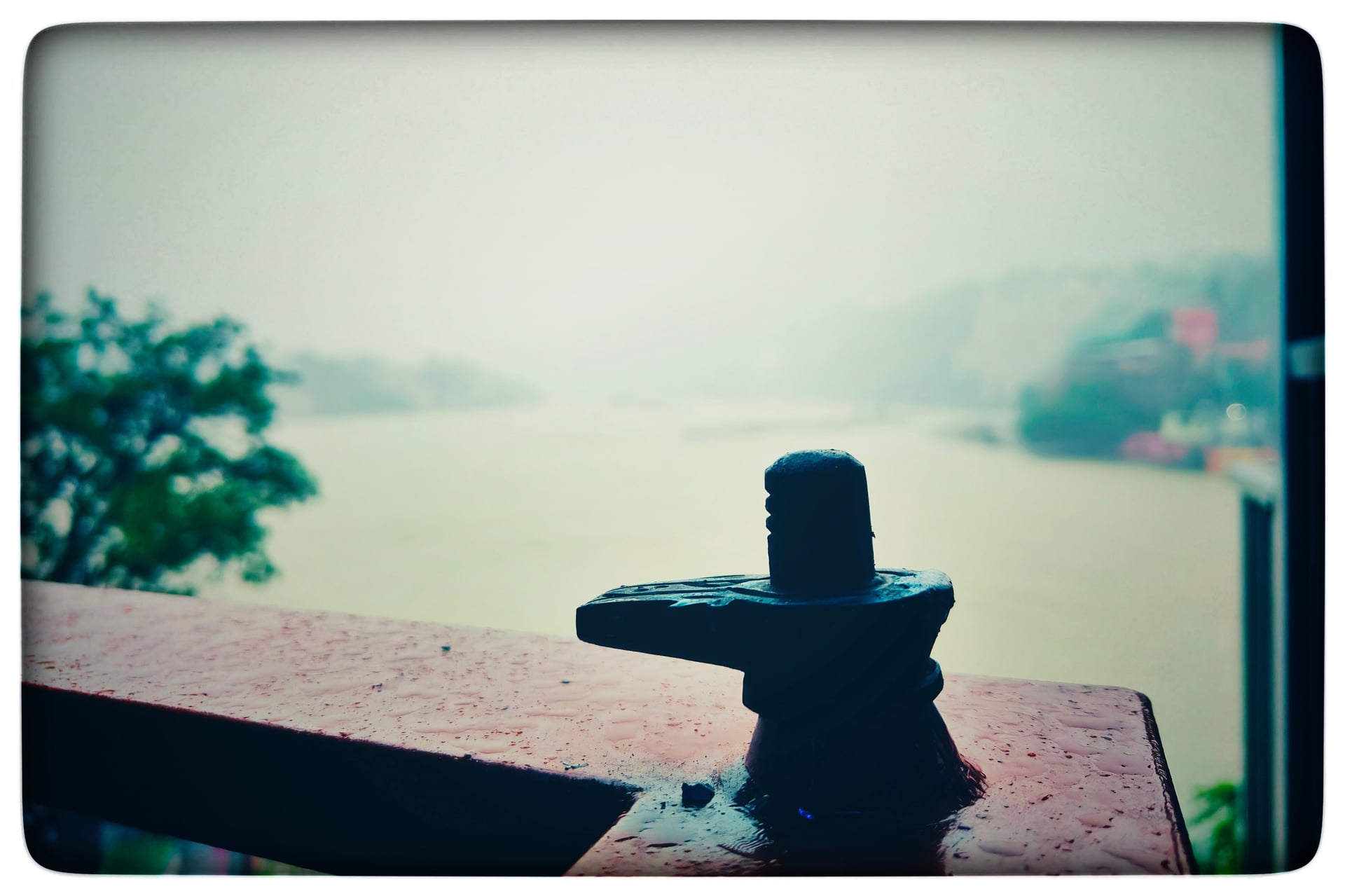 A small black Shiva lingam statue on a wet wooden railing overlooking the hazy Ganga River and distant green hills.