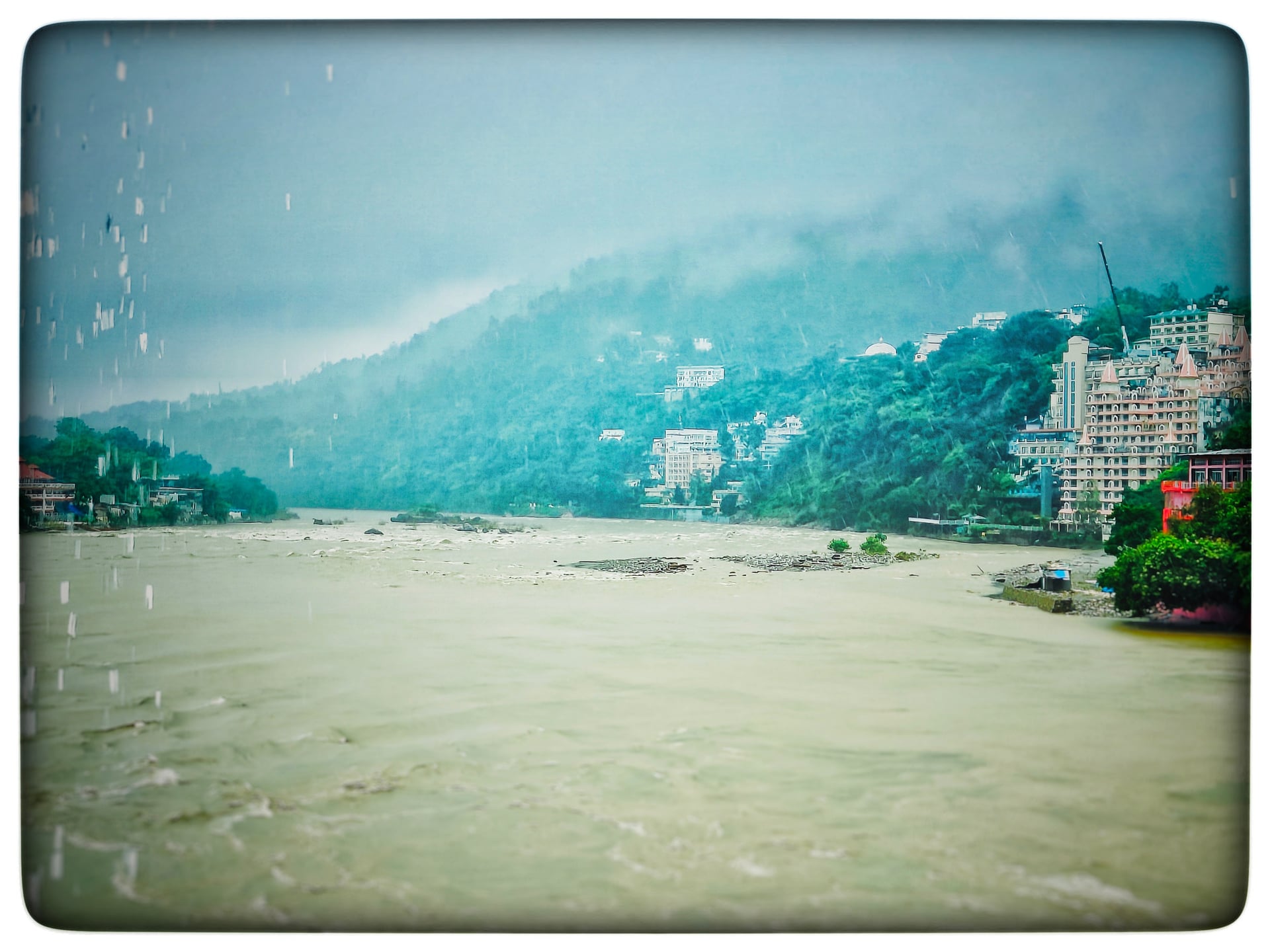 Panoramic view of the wide, muddy Ganges River flowing between lush, misty green hills with buildings lining the banks under a cloudy sky.