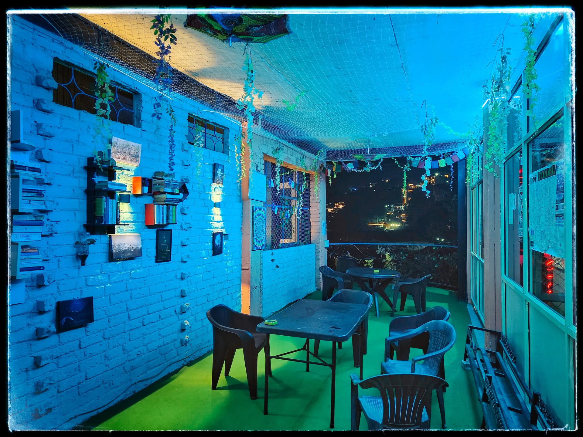 Night view of a hotel terrace with blue lighting, several tables, chairs, and a decorative brick wall. City lights are visible in the distance.