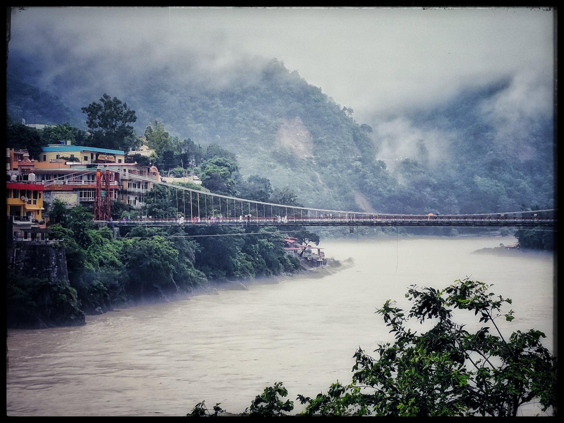 Panoramic view of Laxman Jhula bridge over the misty Ganga River, with colorful buildings on the left bank and fog-shrouded green mountains.