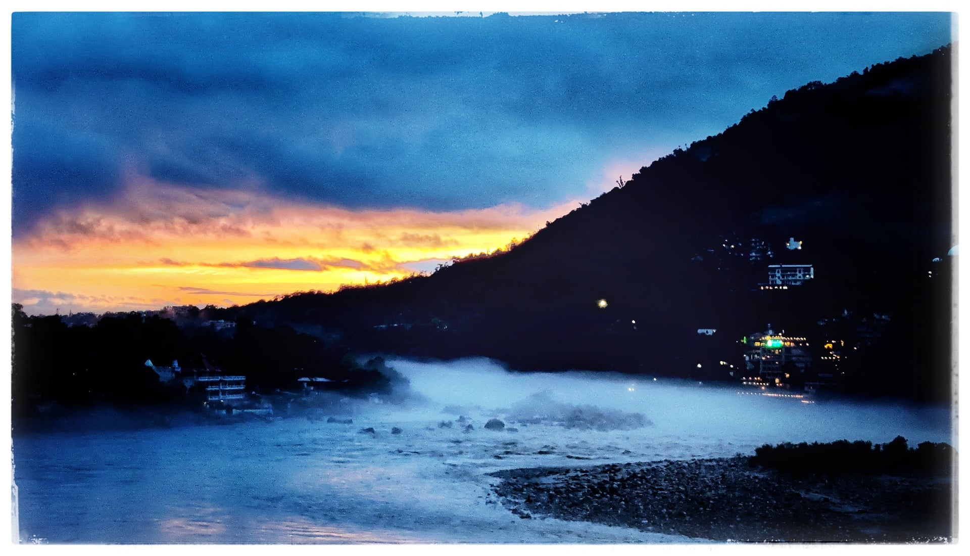 Dramatic sunrise or sunset over the mist-covered Ganga River with a dark forested mountain and distant lights in Rishikesh.