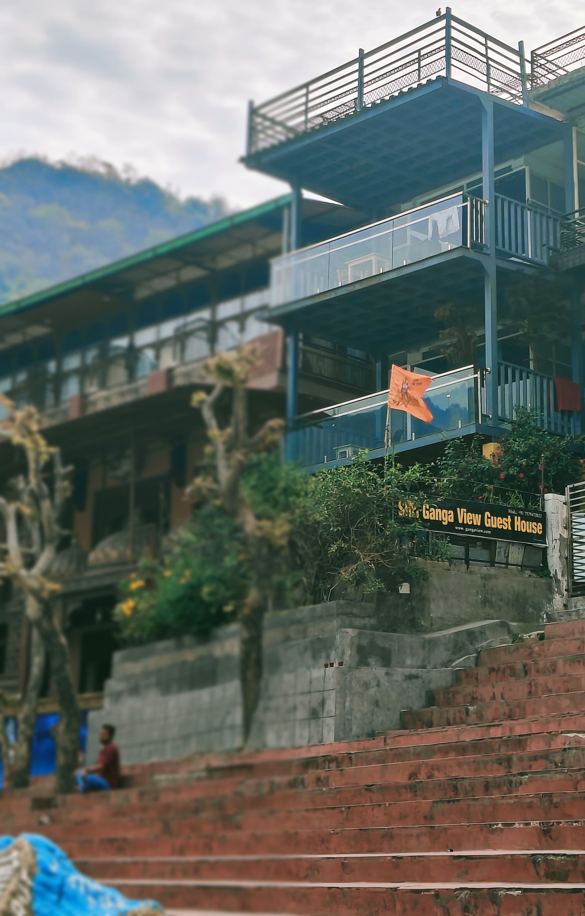 Exterior of Ganga View Guest House, a multi-story building with balconies, steps leading up, and mountains in the background.