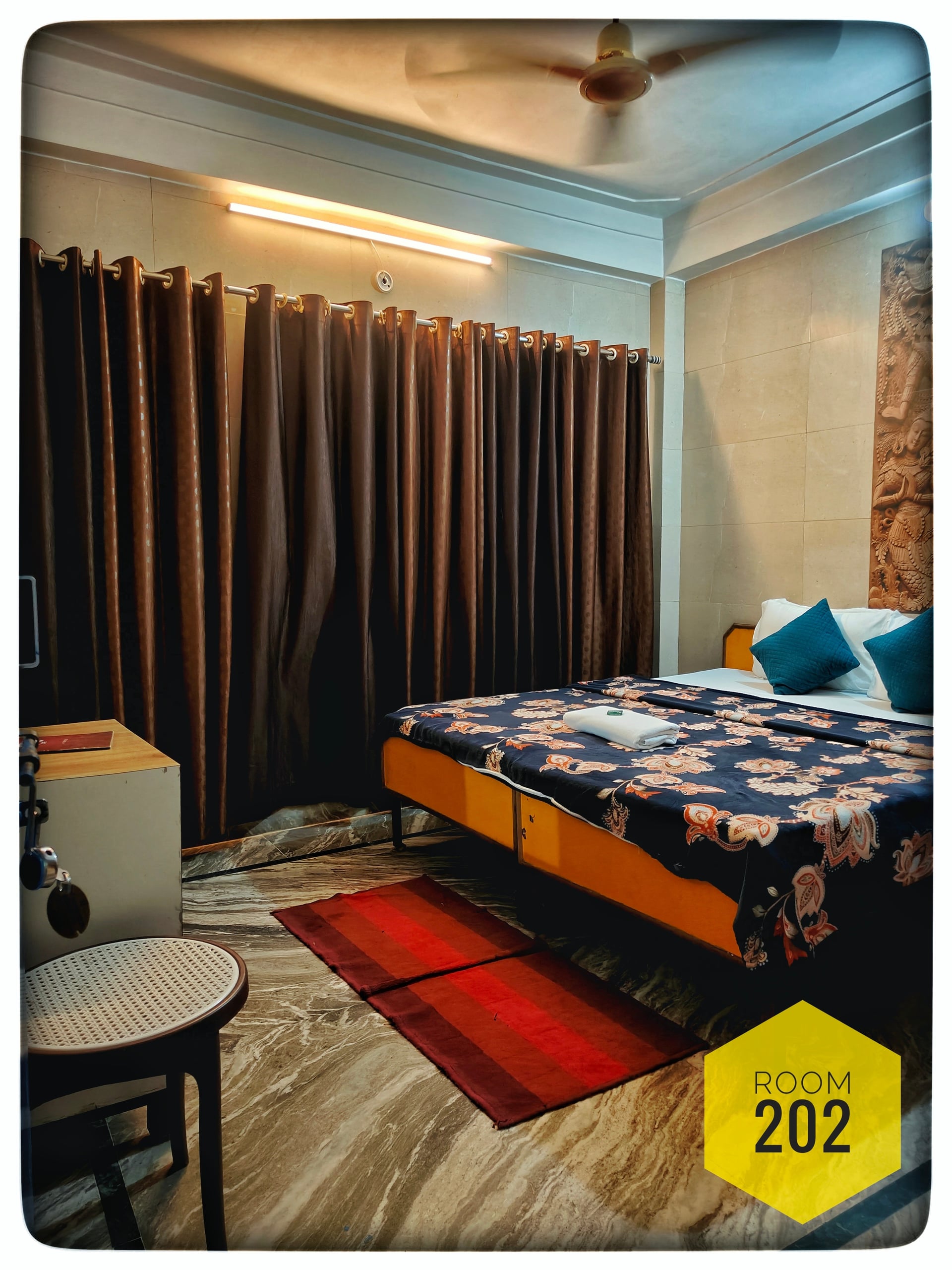 Hotel Ganga View Room 202 featuring a queen bed with floral spread, teal pillows, brown curtains, and a ceiling fan.