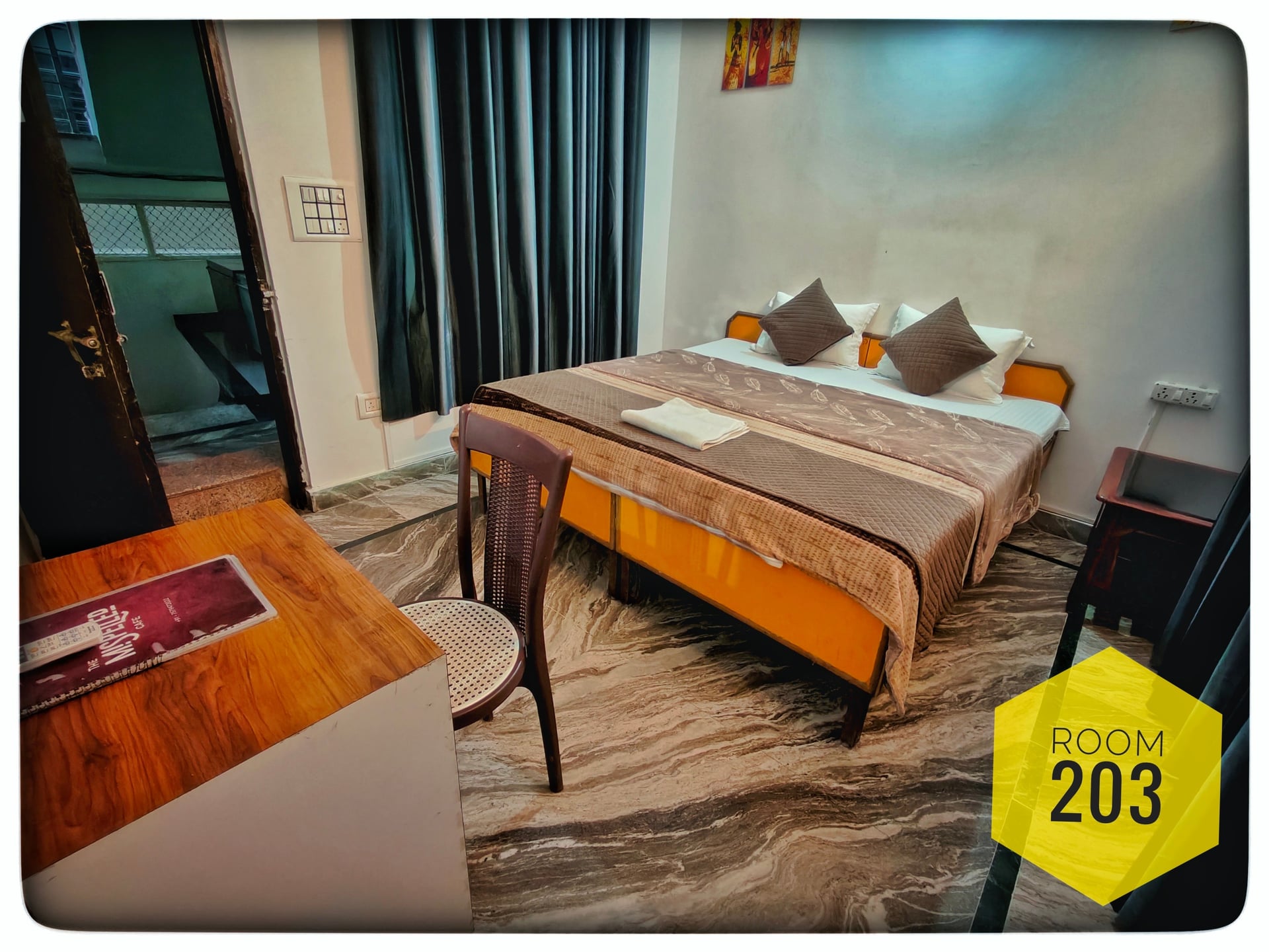 Hotel room 203 with a double bed featuring brown bedding, dark curtains, a small wooden desk, and a chair.