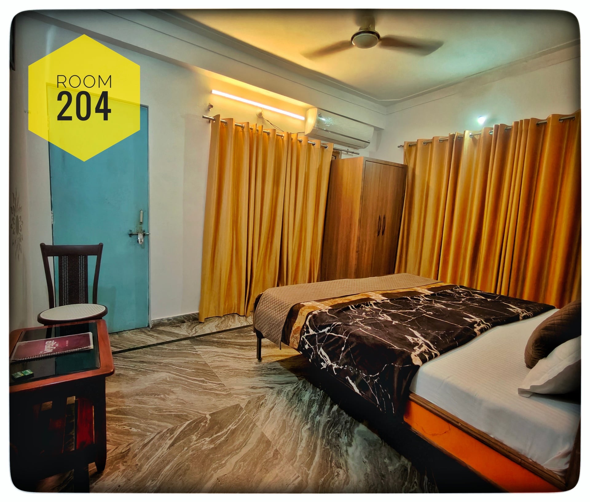 Hotel Ganga View Room 204 with a queen bed featuring a black marble-patterned duvet, wooden wardrobe, and orange curtains.