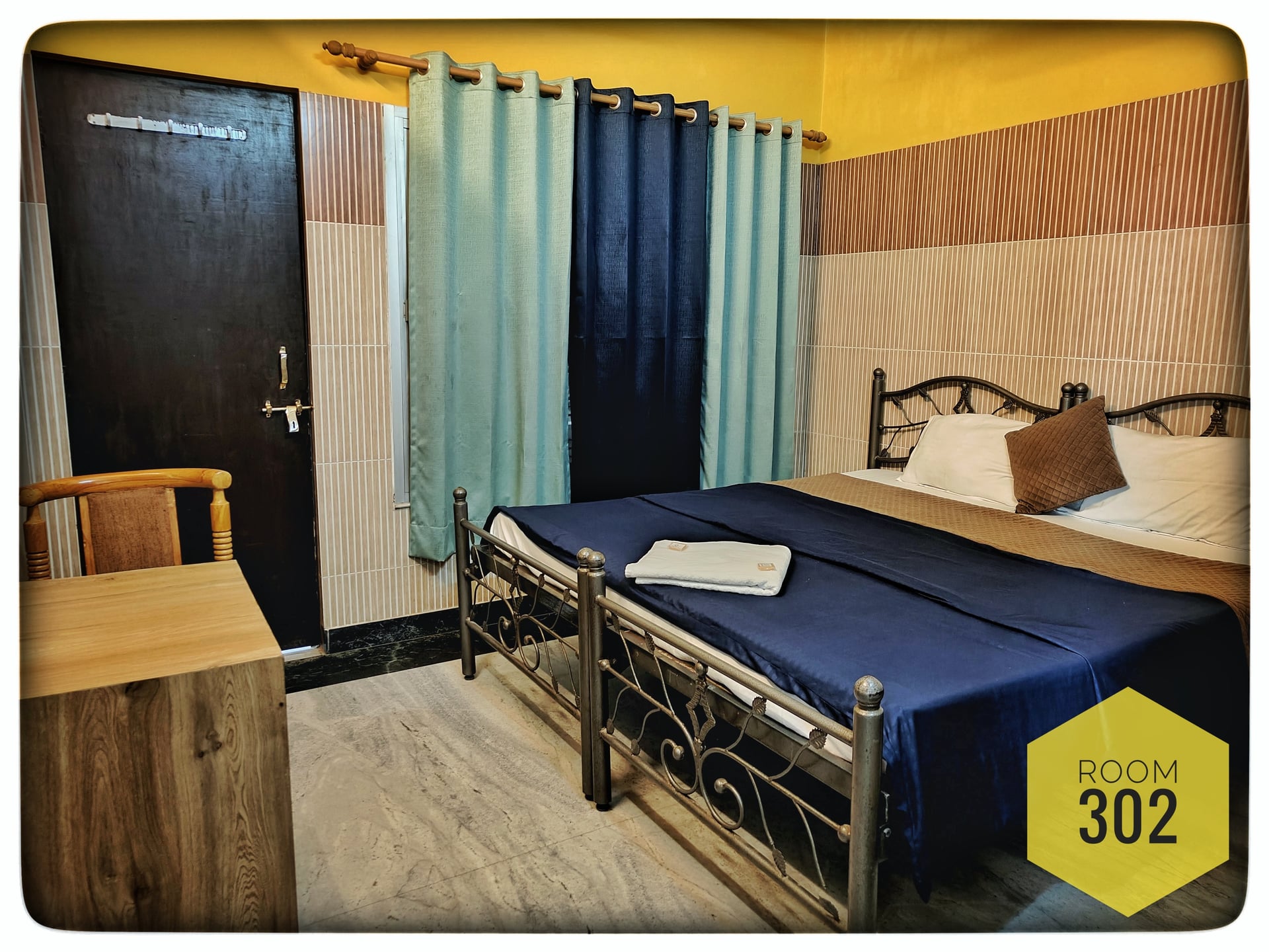 Standard double bedroom with metal-frame bed, blue bedding, wooden desk, and dark door. Yellow walls, tiled sections, blue curtains.