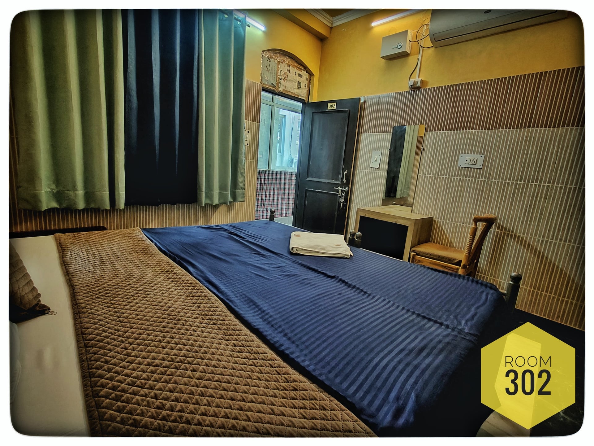 Hotel Ganga View Room 302 with a double bed, dark blue and brown bedding, small dressing table, and chair.