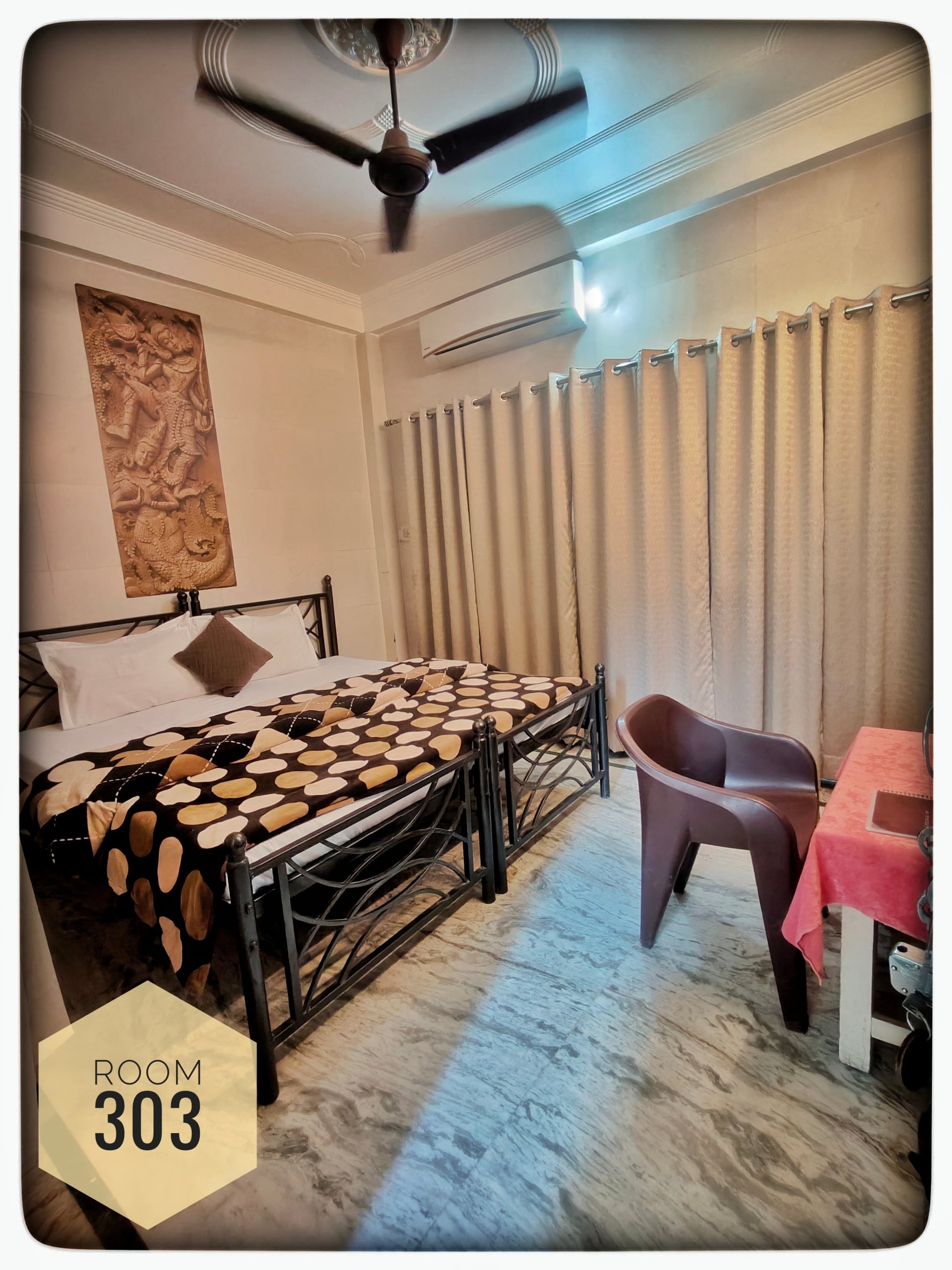 Hotel Ganga View Room 303 featuring two beds with patterned blankets, a ceiling fan, AC, table, and chair.