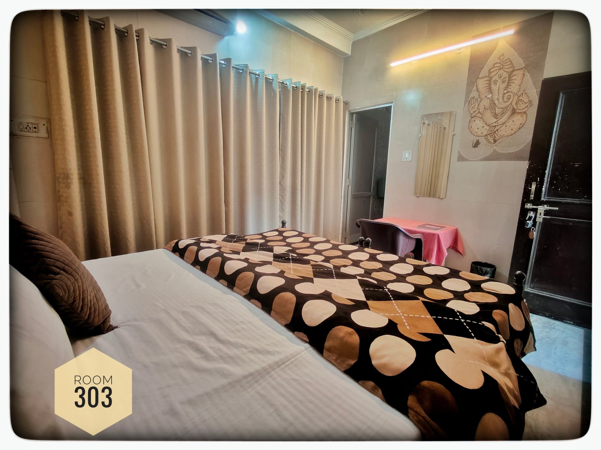 Hotel room 303 with two beds, beige curtains, a Ganesha painting, and a small table.
