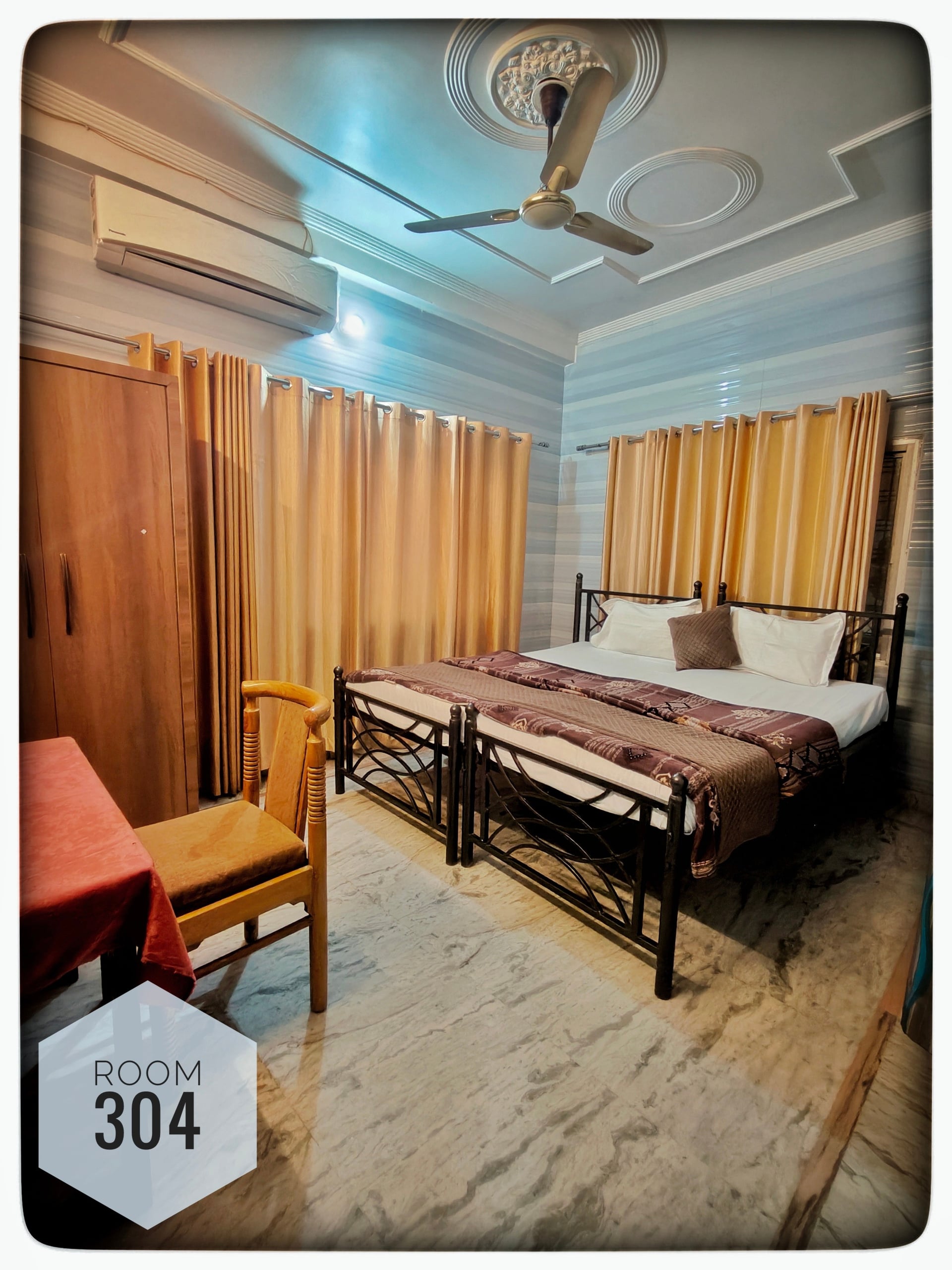 Room 304 with two twin beds, light marble floor, wooden wardrobe, table, chair, golden curtains, air conditioning unit, and ceiling fan.