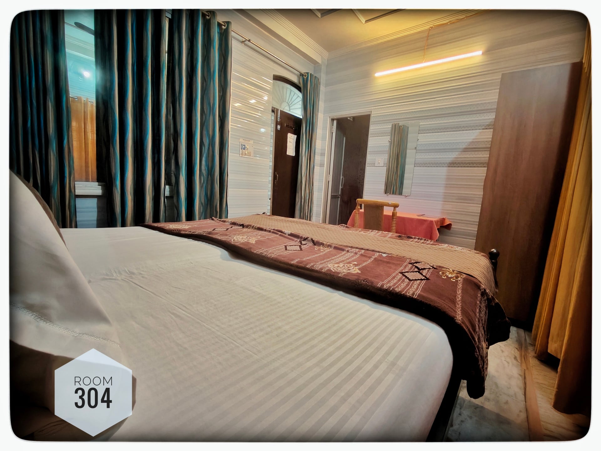 Hotel Room 304 with a king-size bed, blue curtains, wooden wardrobe, and a small dining table with chairs. Room number visible.
