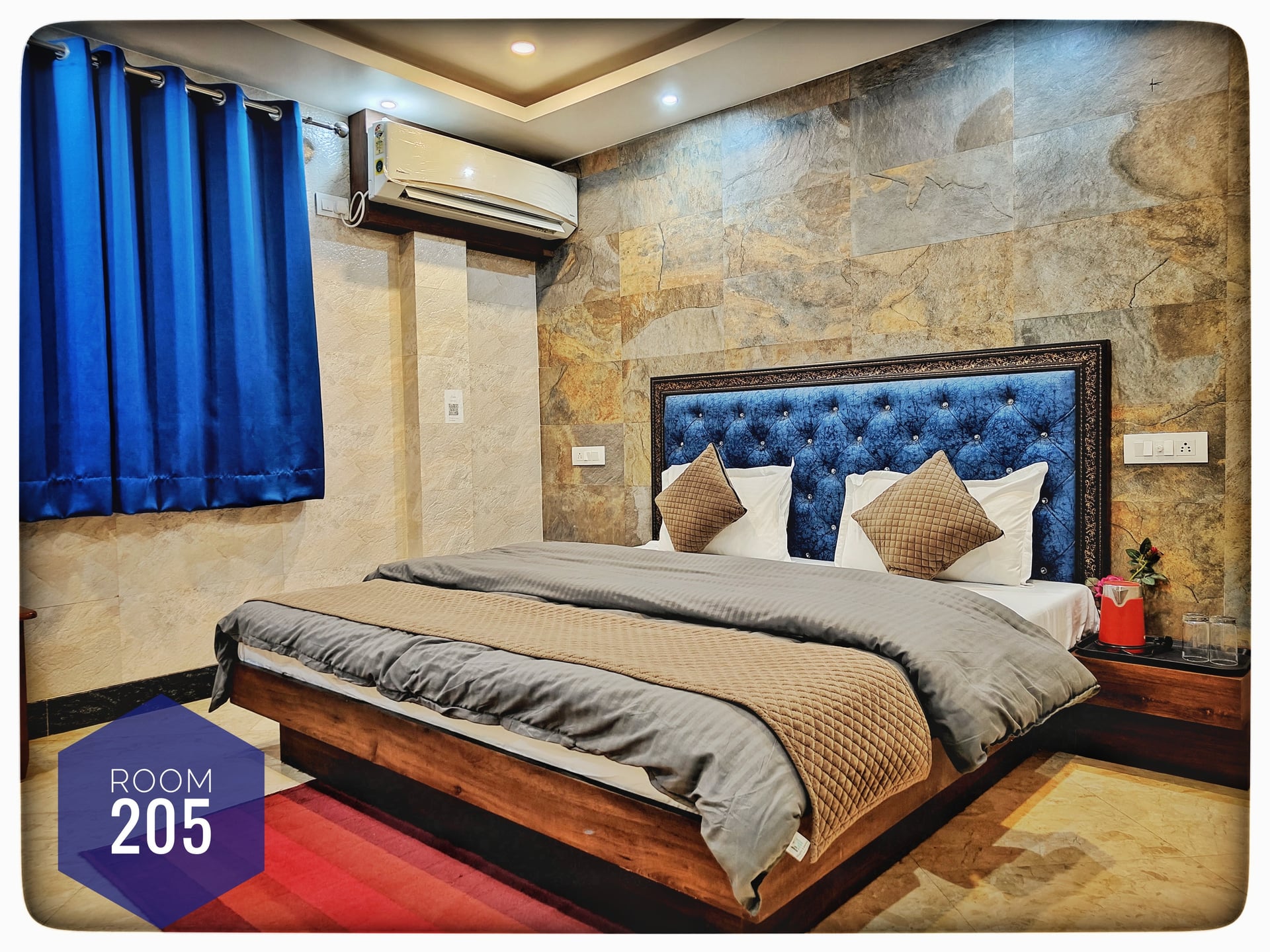 A modern hotel bedroom with a king-size bed, blue tufted headboard, grey bedding, blue curtains, and stone-textured wall.