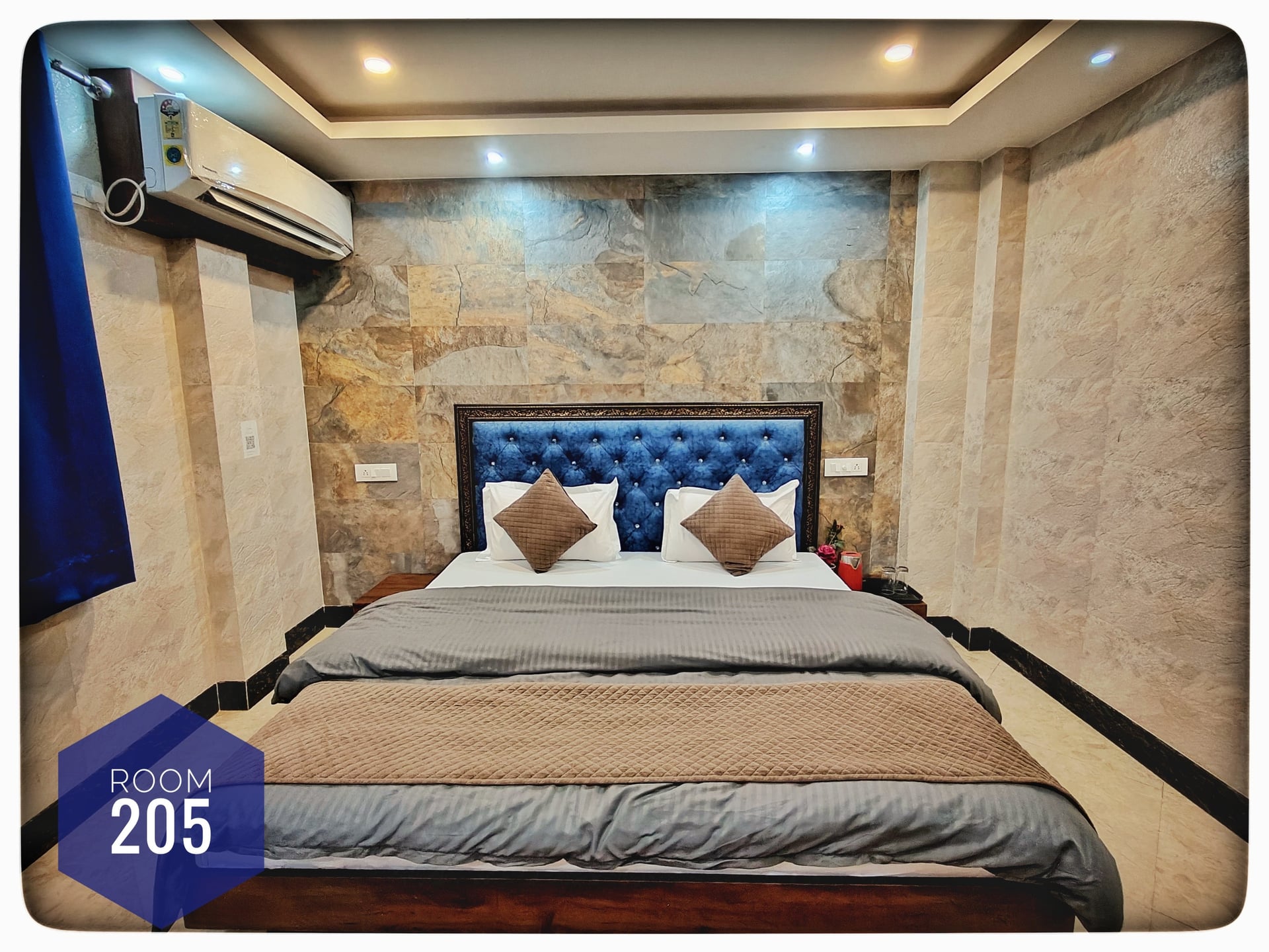 Hotel Room 205 with a large bed, blue tufted headboard, stone accent wall, air conditioner, and blue curtains.