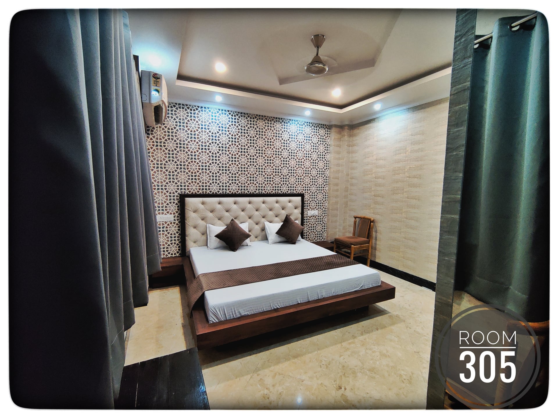 Modern hotel bedroom with king-size bed, tufted headboard, patterned wall, ceiling fan, and accent chair. Room 305.