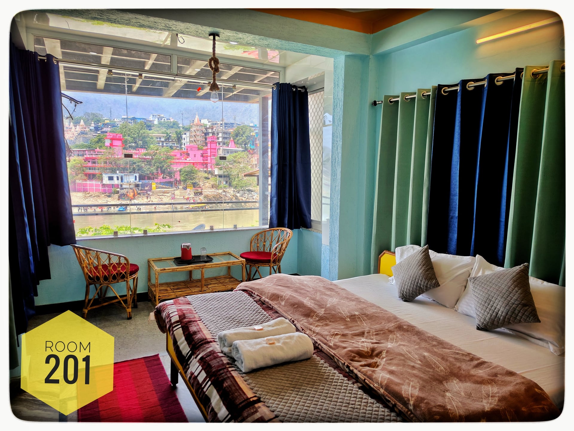 Room 201 at Hotel Ganga View with a cozy bed, seating area, and panoramic window offering views of the Ganga River and pink temples.