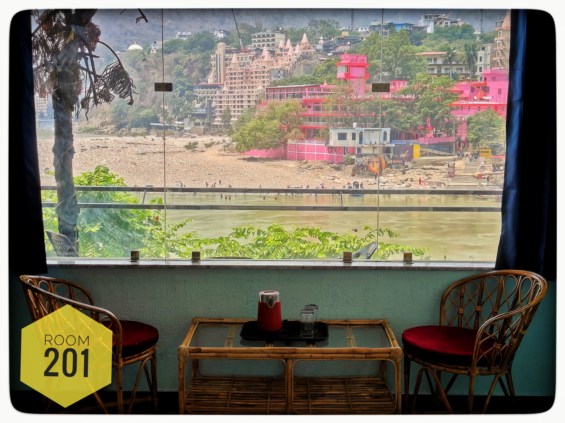 A room view of the Ganga river from Hotel Ganga View, Rishikesh. Two rattan chairs and a table are in the foreground, facing the river and colorful buildings on the far bank.