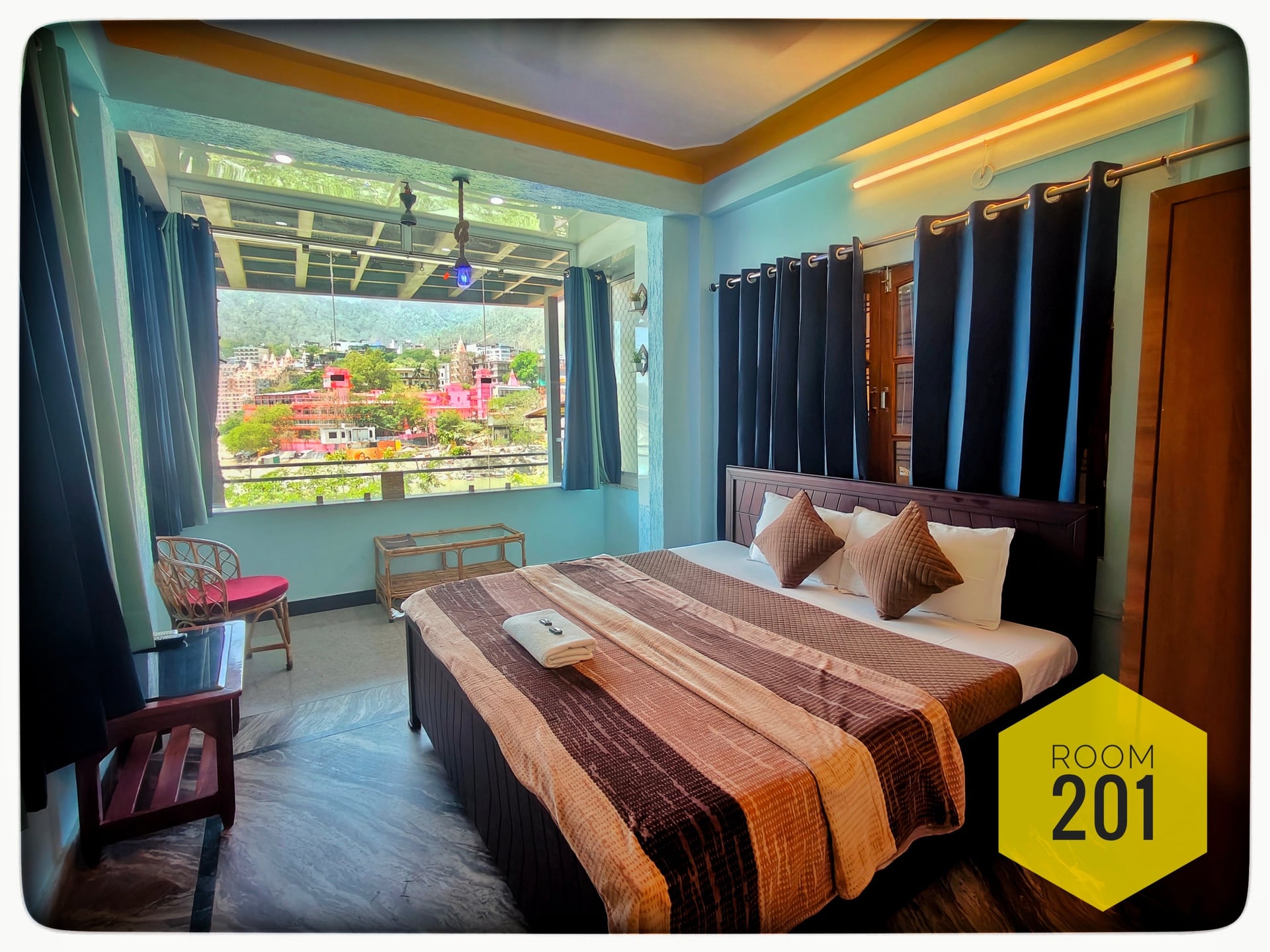 Hotel Ganga View Room 201 with a queen bed, sitting area, and large window overlooking colorful buildings and hills.