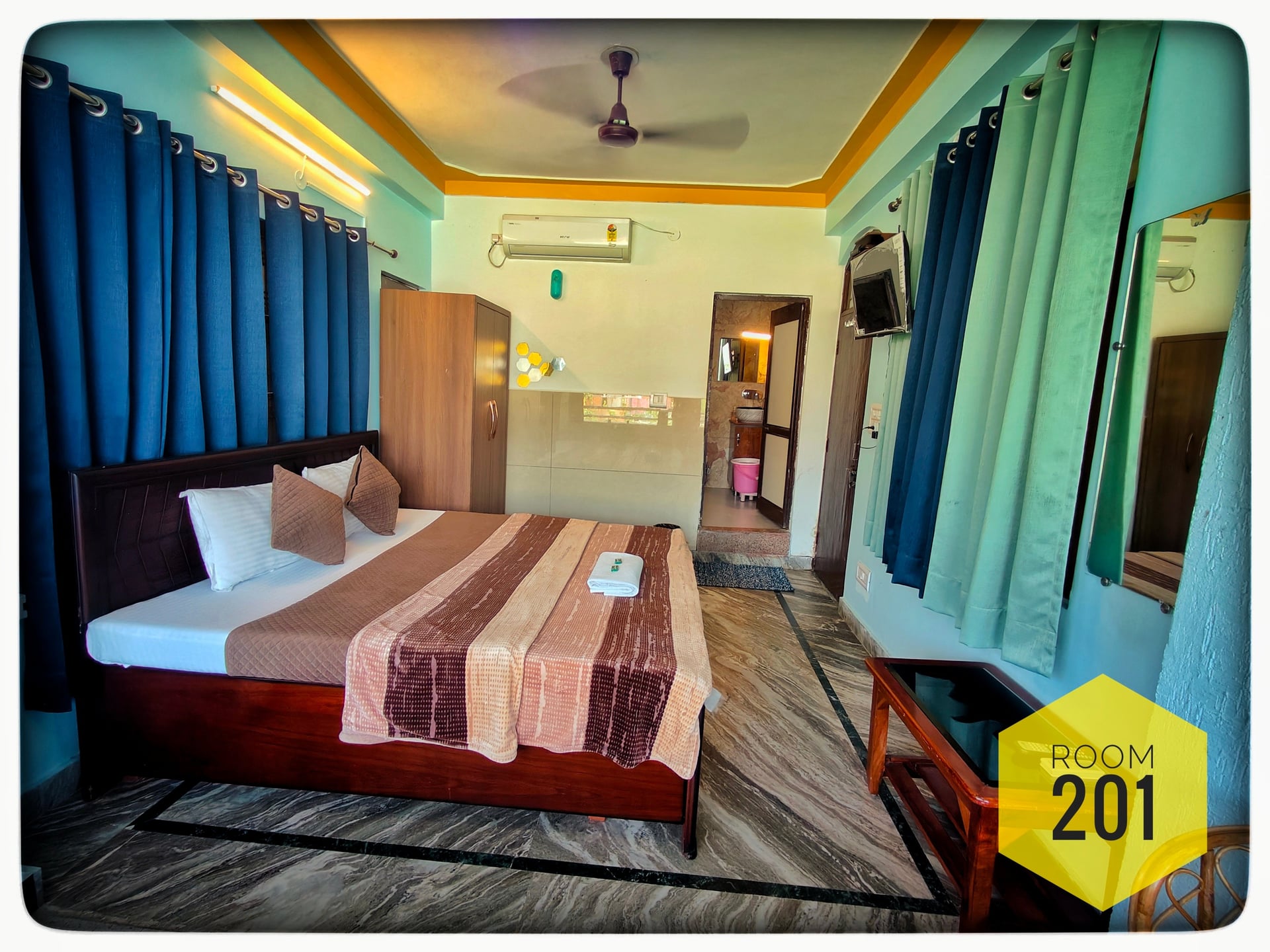 Hotel room 201 with a double bed, dark wooden furniture, blue curtains, and a partial view of the bathroom.