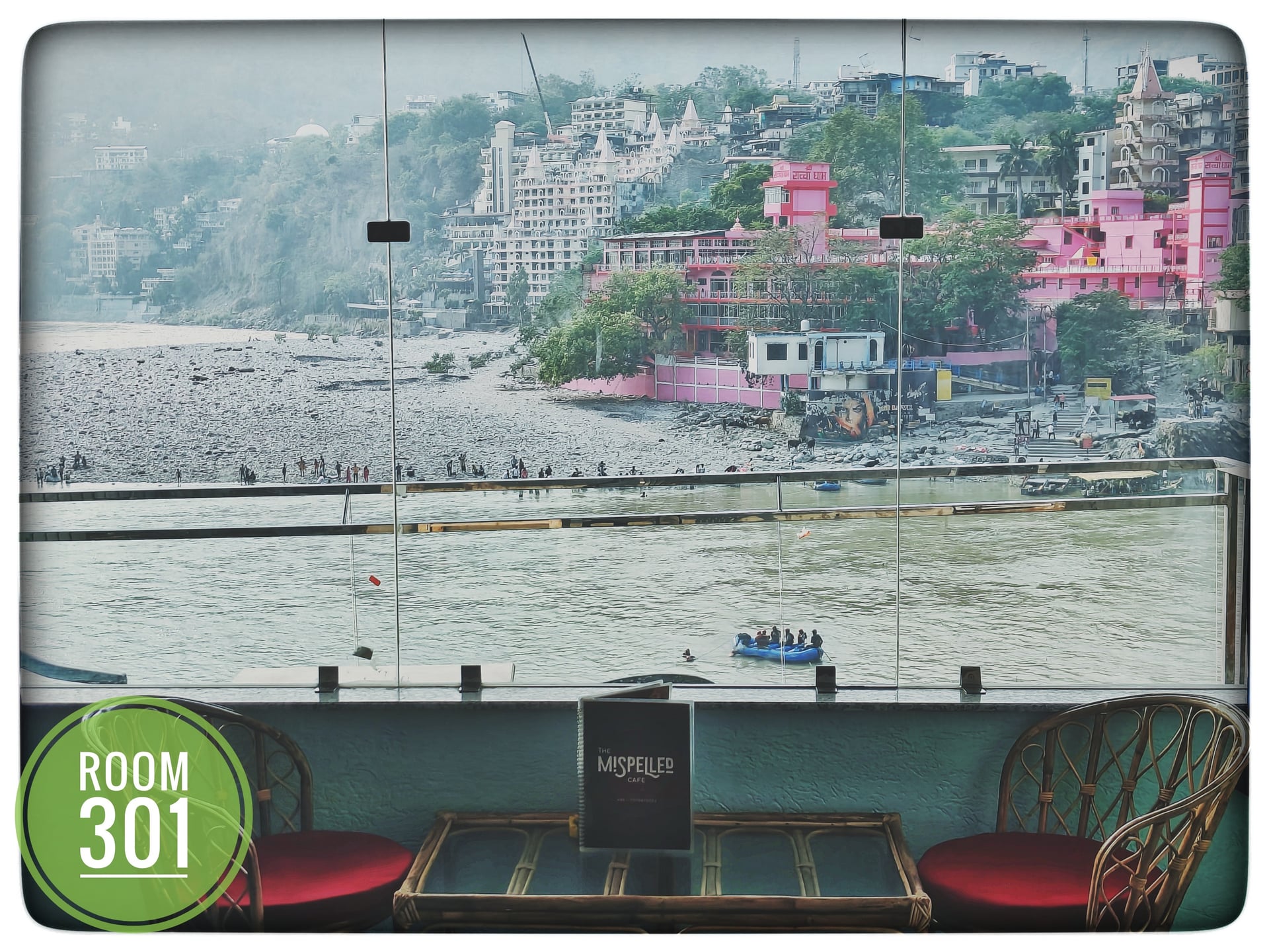 View from hotel room 301 overlooking the Ganga River with buildings on a hill, a raft, and two indoor chairs.