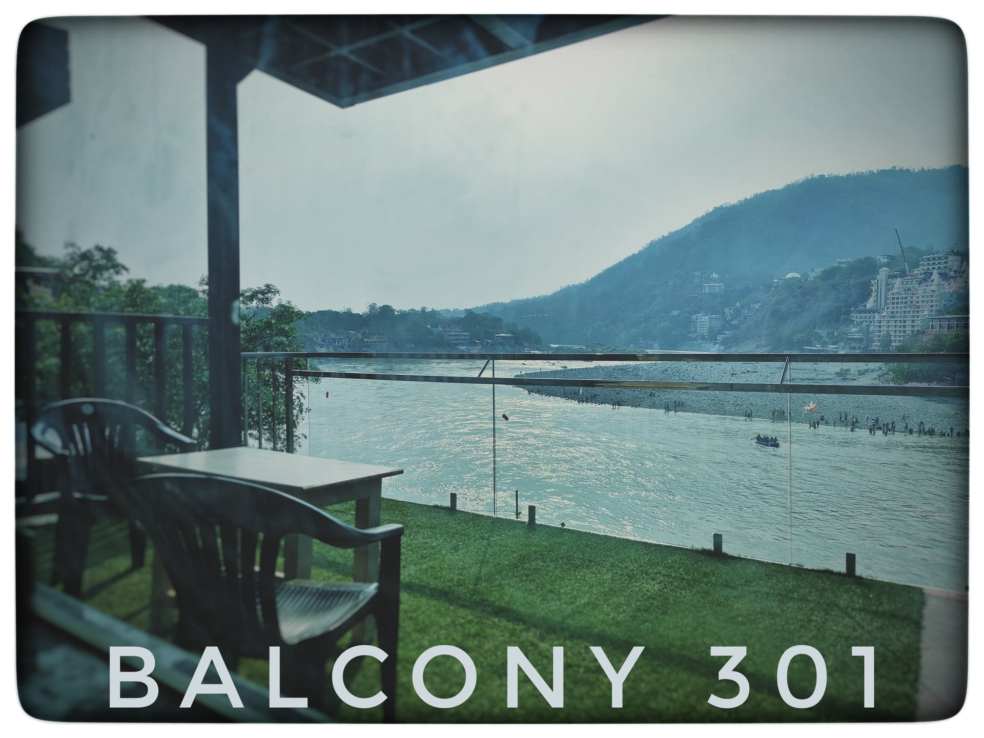Balcony of room 301 with a small table and two chairs, offering a scenic view of the wide Ganga River and distant mountains.