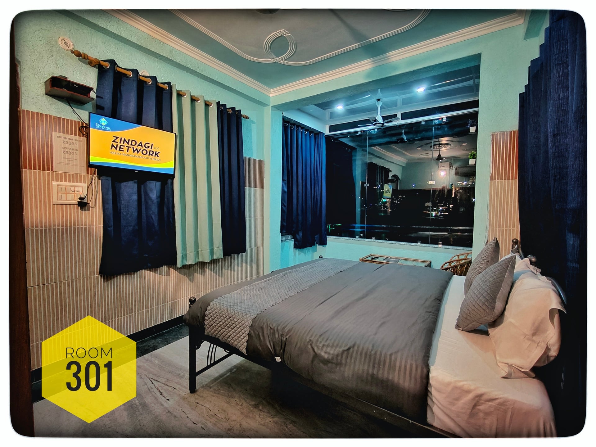 Hotel Ganga View Room 301 with a comfortable bed, grey and white linens, a wall-mounted TV, and a large window.