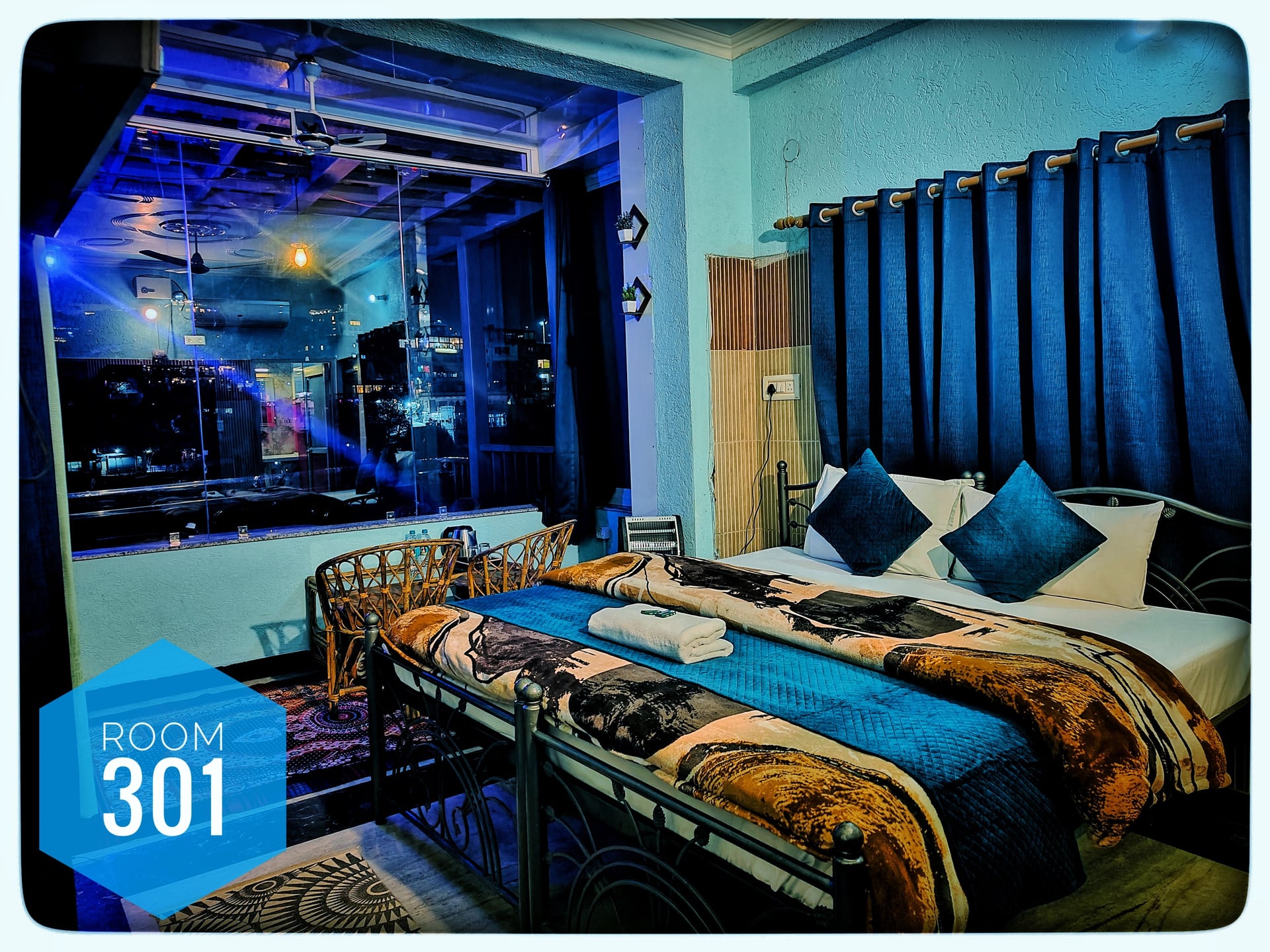 Hotel Ganga View's Room 301 with a king-size bed, blue bedding, and a large window overlooking a city night view.