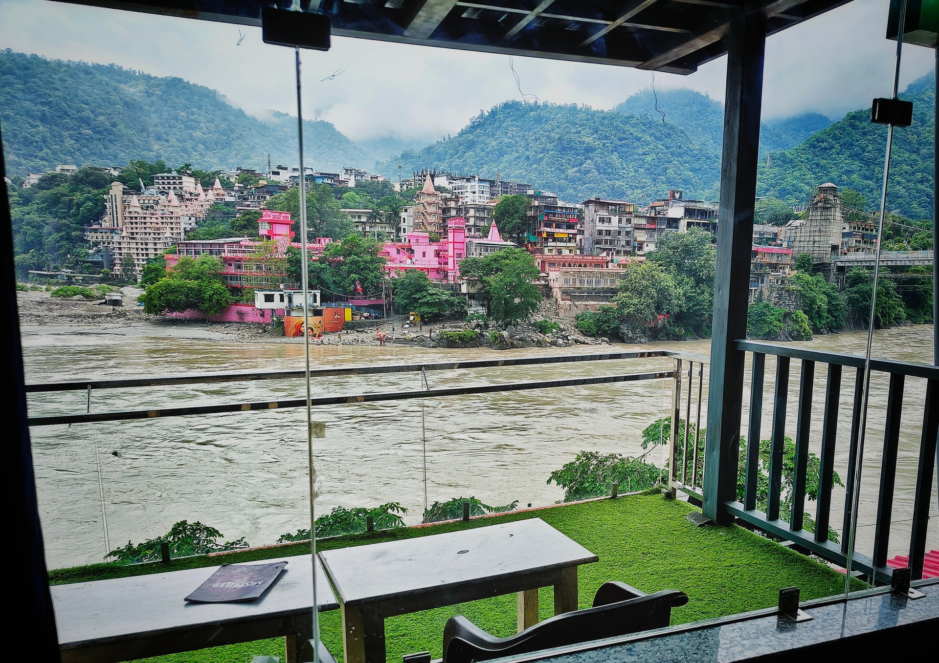View from hotel balcony showing Ganges River, colourful Rishikesh buildings, and lush green mountains under a cloudy sky.