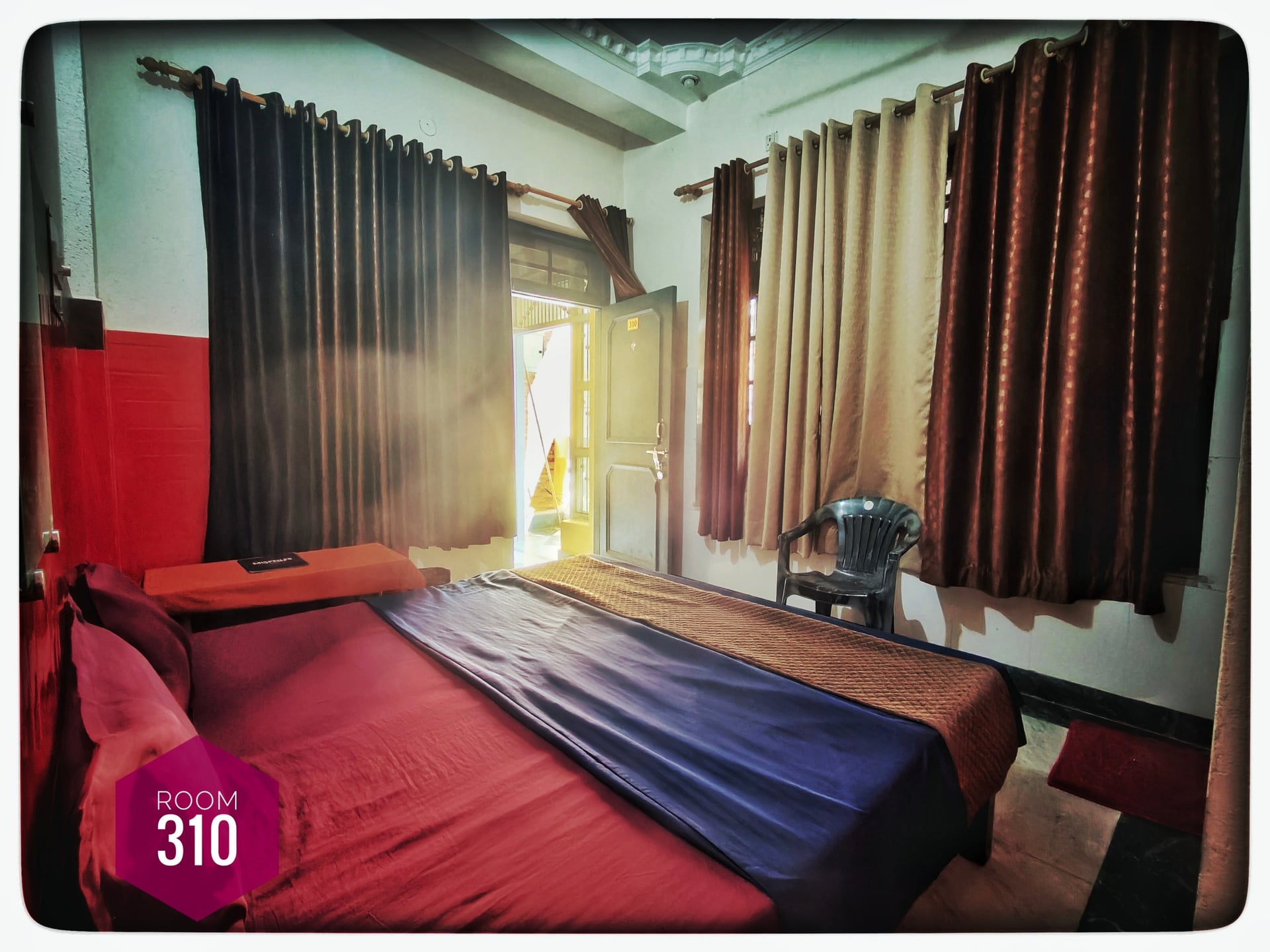 Hotel Ganga View, Rishikesh. Room 310 with a bed, colorful linens, curtains, and an open door letting in bright daylight.