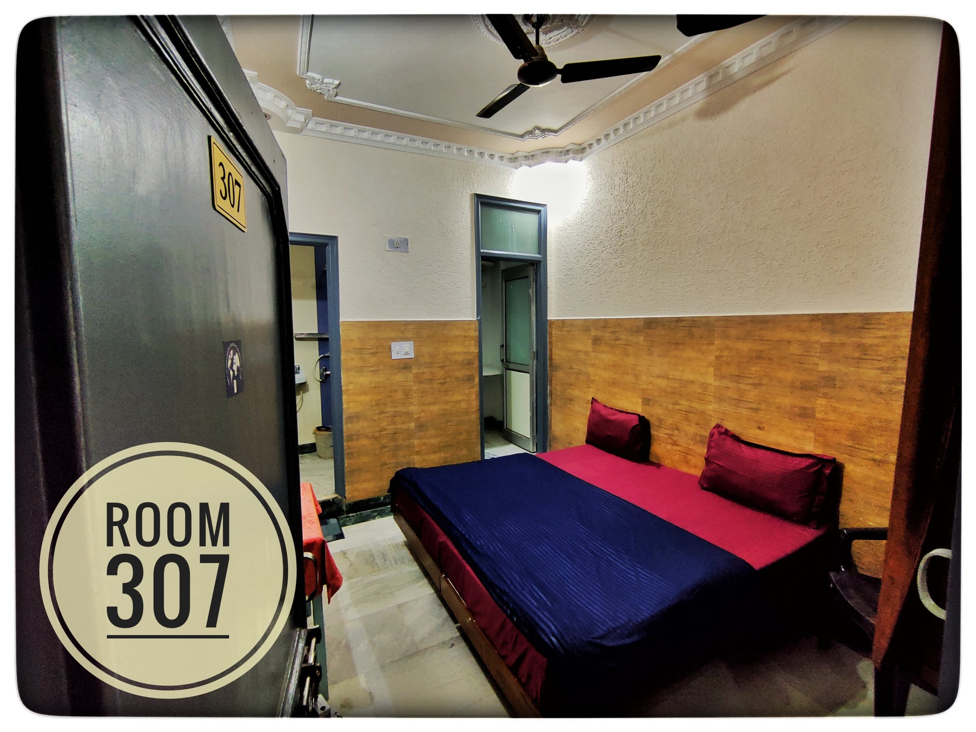 View of Room 307 at Hotel Ganga View, featuring a bed with red and blue linens, wood-paneled walls, and a ceiling fan.