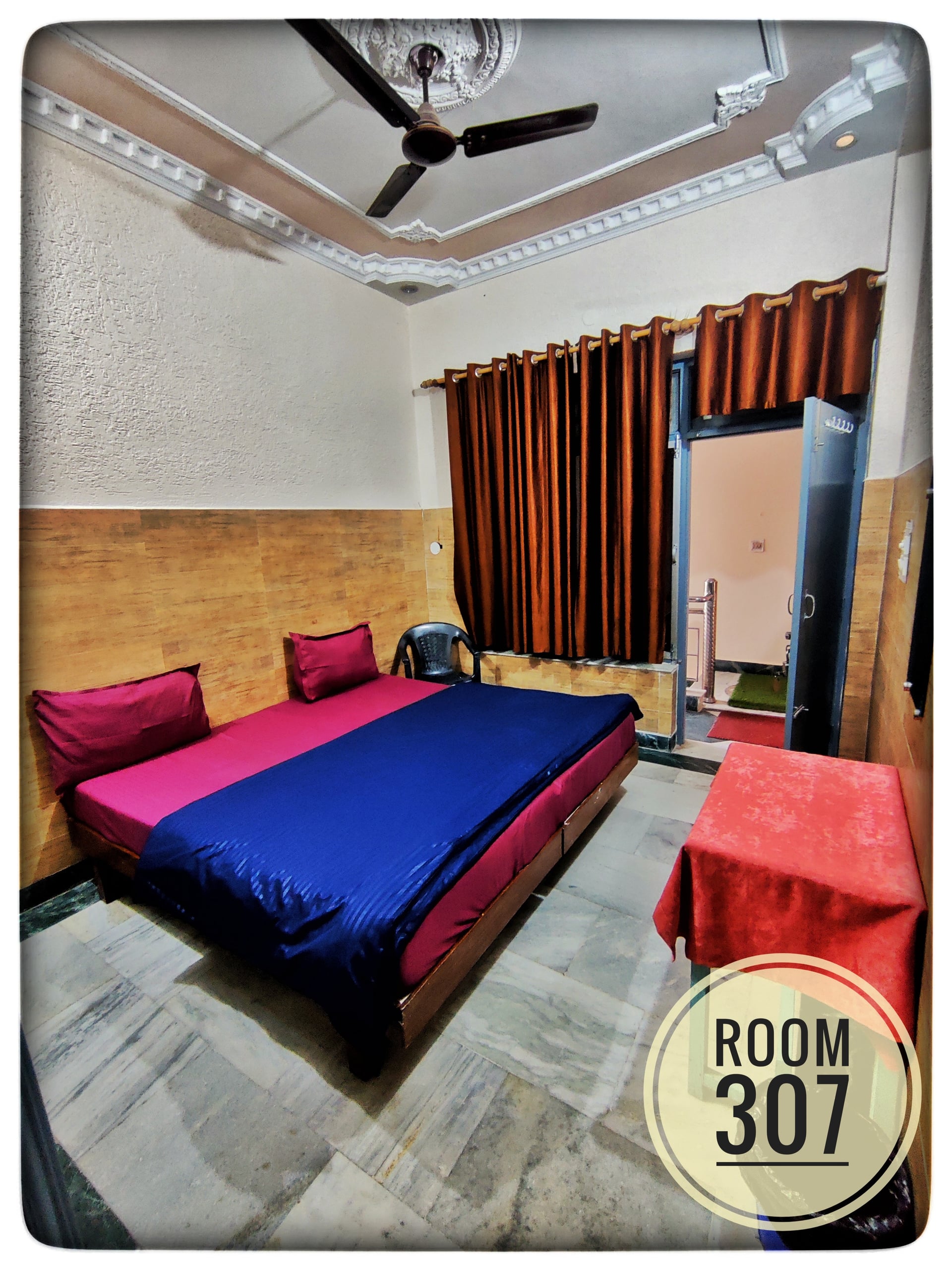 Bedroom 307 with a double bed, pink and blue bedding, a small table with a red cloth, and a ceiling fan.
