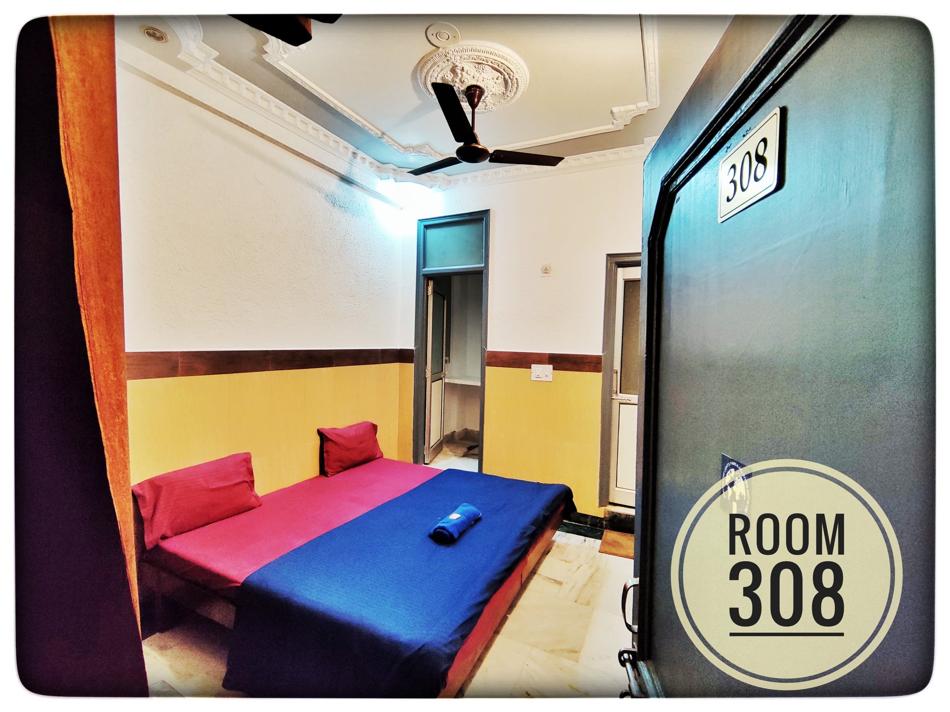 Standard Room 308 at Hotel Ganga View. Features a twin bed with red and blue linens, ceiling fan, and yellow and white walls.
