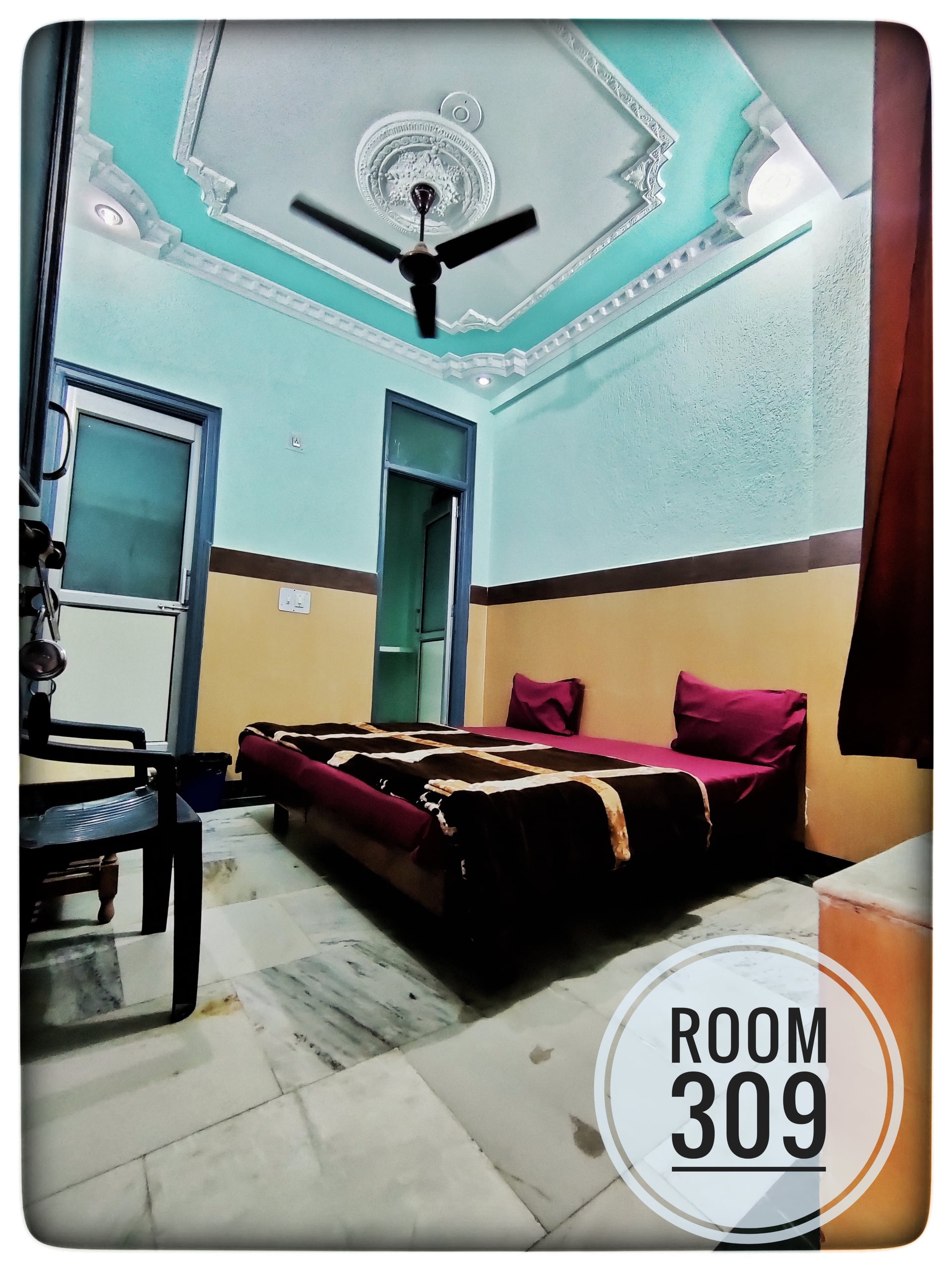 Standard double bedroom at Hotel Ganga View, featuring a bed with maroon linens, light blue walls, and a ceiling fan.